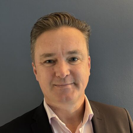 Een man met kort haar, gekleed in een donker colbert en een lichtroze overhemd, staat voor een effen donkergrijze achtergrond.