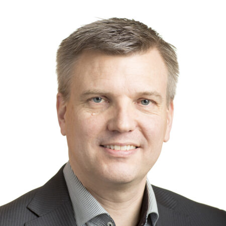 Een man van middelbare leeftijd met kort grijs haar, gekleed in een donker krijtstreeppak en een licht gestreept overhemd, en glimlachend naar de camera tegen een effen witte achtergrond.