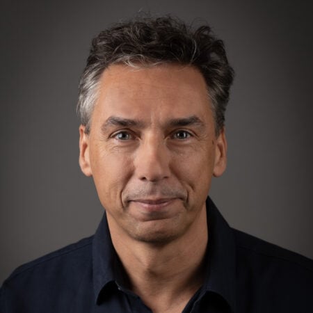 Een man van middelbare leeftijd met kort, grijzend haar, gekleed in een donker overhemd met knopen, poseert voor een portret tegen een donkere achtergrond.