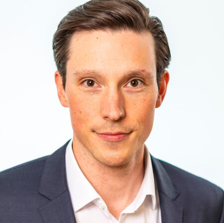 Een man met kort bruin haar, gekleed in een wit overhemd en een donker colbert, kijkt rechtstreeks in de camera tegen een effen witte achtergrond.