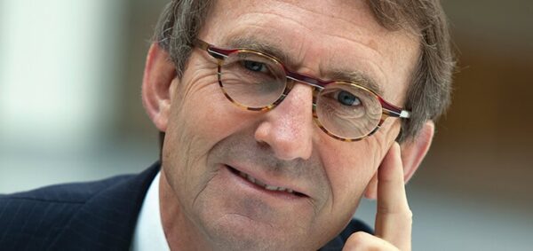 Een man van middelbare leeftijd met kort haar en een bril, gekleed in een pak, kijkt met een lichte glimlach in de camera en legt zijn wang op zijn hand.