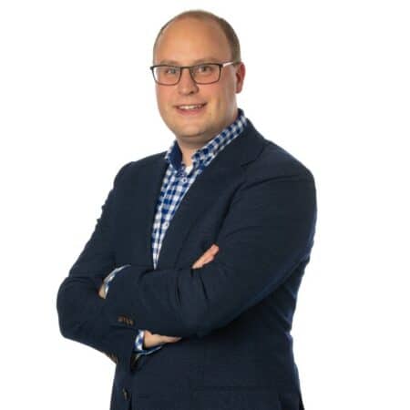 Een man met een bril, gekleed in een marineblauw jasje en een blauw geruit overhemd, staat met gekruiste armen tegen een witte achtergrond.