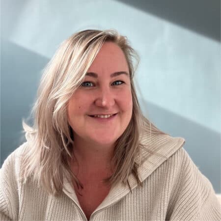 Een vrouw met blond haar en een witte trui glimlacht naar de camera. Ze zit tegen een lichtblauwe achtergrond.