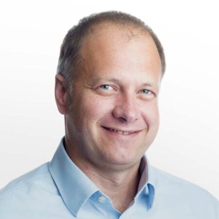 Een man van middelbare leeftijd met kort haar en een terugtrekkende haarlijn glimlacht, gekleed in een lichtblauw overhemd tegen een witte achtergrond.