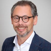 Een man met een bril, grijzend haar en een baard lacht op een portret. Hij draagt een blauwe blazer en een wit shirt.