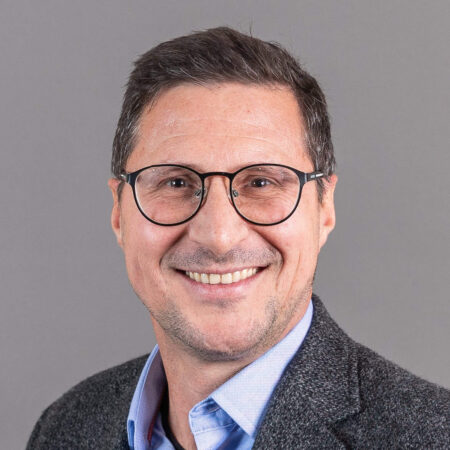 Een man met kort donker haar, een bril en een grijs jasje over een blauw shirt, lacht tegen een grijze achtergrond.