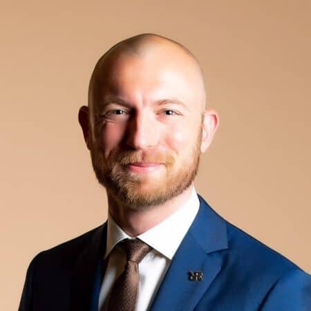 Een kale man met een baard, gekleed in een blauw pak en een bruine stropdas, glimlacht tegen een beige achtergrond.