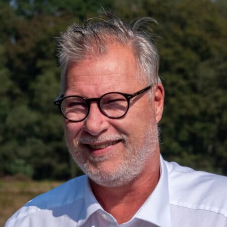 Een man met een bril en een baard staat glimlachend buiten voor de bomen, gekleed in een wit shirt.