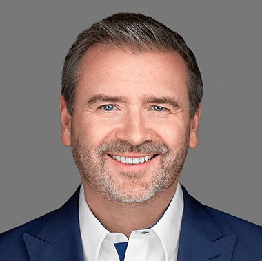 Man met kort haar en een baard, gekleed in een blauw pak en een wit overhemd, glimlachend voor een grijze achtergrond.