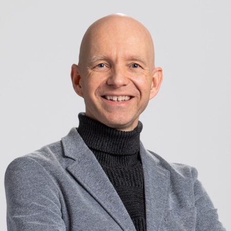 Een lachende kale man in een grijs jasje en een zwarte coltrui poseert tegen een effen achtergrond.