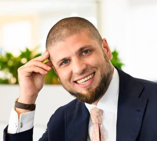 Een man in pak en stropdas lacht, met zijn hand op zijn slaap. Binnenomgeving met wazige planten op de achtergrond.