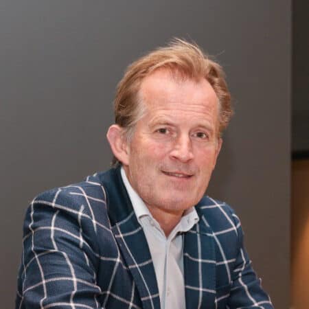 Een persoon in een geruit jasje en een wit overhemd lacht naar de camera tegen een effen achtergrond.