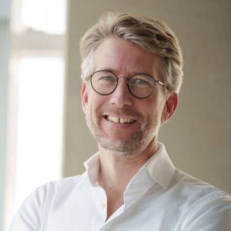 Een man met een bril en een baard, gekleed in een wit overhemd, glimlacht terwijl hij binnen staat.