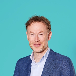 Man met kort haar en een baard, gekleed in een blauw pak en een wit overhemd, staande tegen een effen blauwgroene achtergrond.