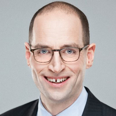Een kale man met een bril lacht naar de camera. Hij draagt een donker pak en een lichtblauw overhemd.