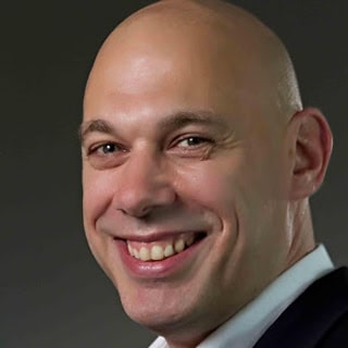 Een persoon met een kaal hoofd, glimlachend, gekleed in een donker jasje en een wit overhemd, tegen een neutrale achtergrond.