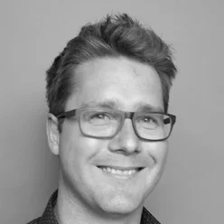 Een man met een bril en kort haar lacht op een zwart-witportret.