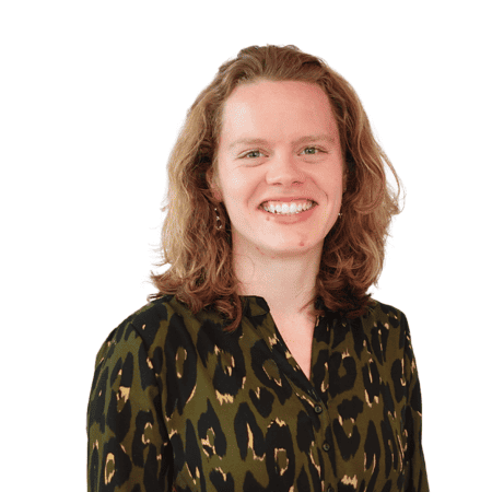Een persoon met schouderlang krullend haar lacht naar de camera. Ze dragen een donkergroene blouse met luipaardprint tegen een effen witte achtergrond.