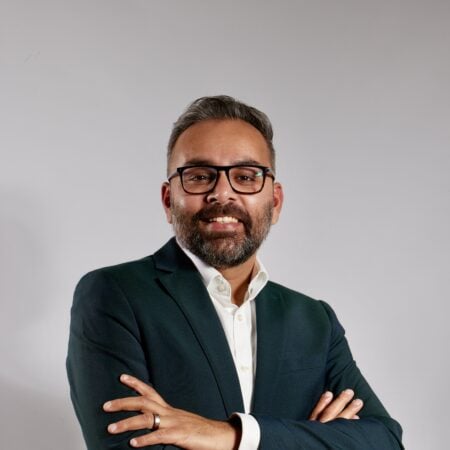 Een man met kort haar, een bril en een baard, gekleed in een donker colbert en een wit overhemd, staat met gekruiste armen tegen een effen, lichte achtergrond.