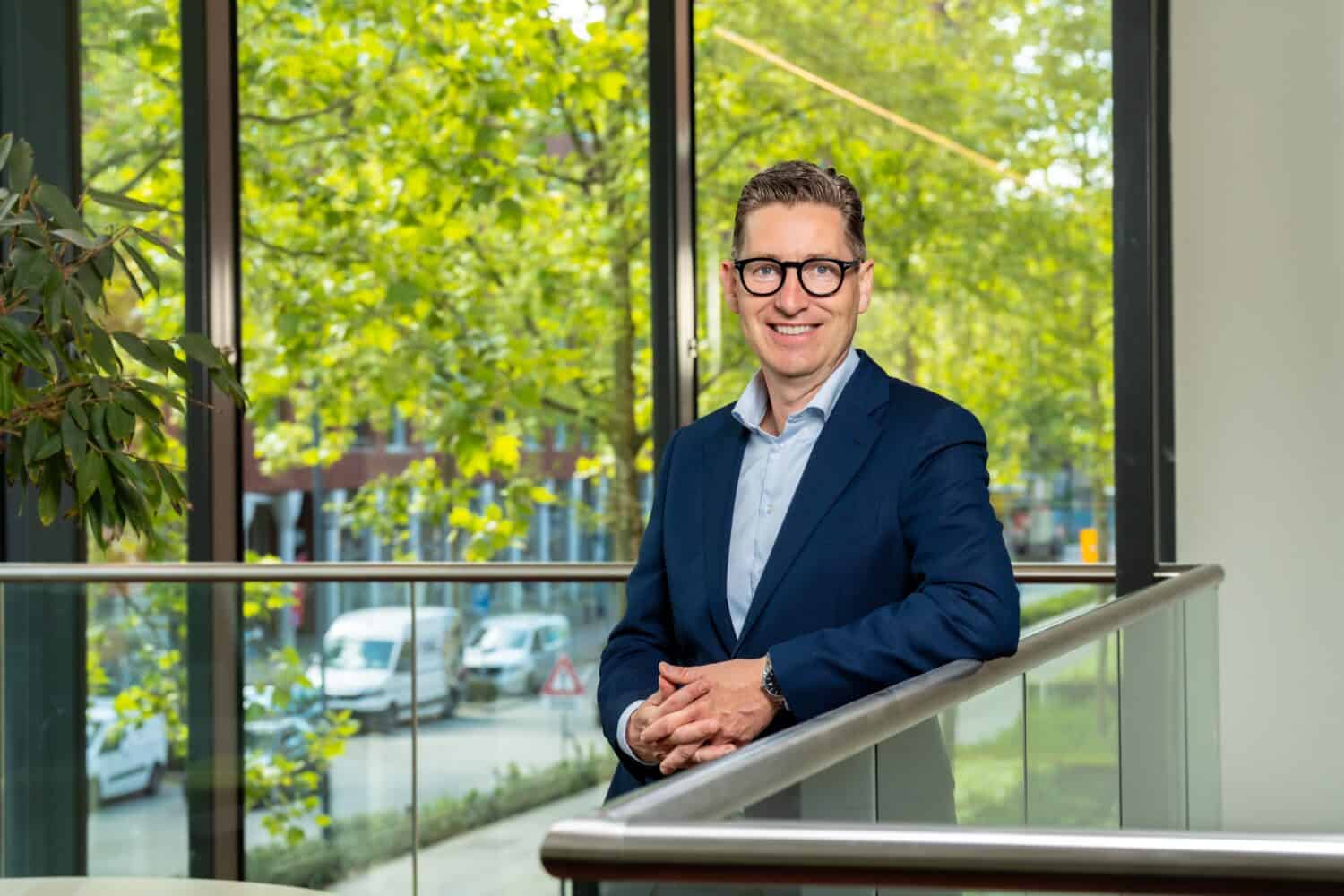 Rick van der Kleij Een man in een blauw pak en een bril staat binnen bij de balustrade en lacht. Op de achtergrond zijn grote ramen en groene bomen te zien.