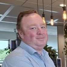 Een man met kort lichtbruin haar en een lichtblauw shirt staat binnen bij hangende lampen en planten en kijkt iets naar links.