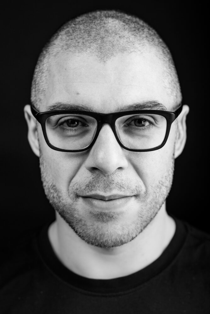 DavideCeolin Een man met een kaalgeschoren hoofd, bril en lichte stoppelbaard kijkt recht in de camera tegen een effen donkere achtergrond en denkt na over meertalige AI-bias in bedrijfsapplicaties.