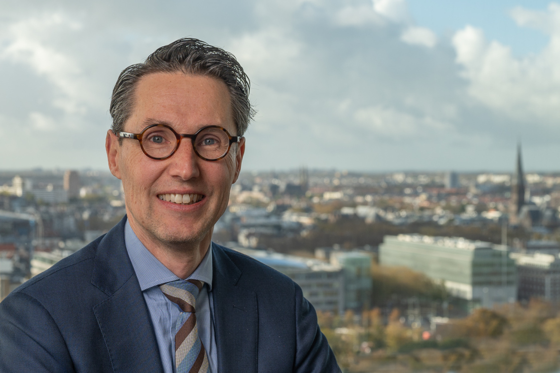 Frits Grotenhuis, directeur Topsector ICT Een man in pak en stropdas met een bril lacht naar de camera, met een stadsgezicht en een bewolkte lucht op de achtergrond.