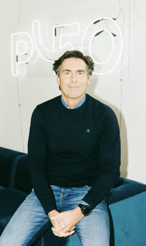 Raymond Hüner Een man in een marineblauwe trui en spijkerbroek zit op een blauwe bank voor een neonreclame met de tekst 'PLUG'.