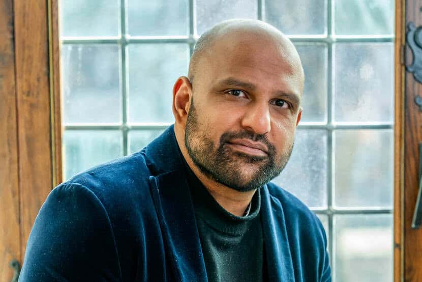 Haroon Sheikh – sr scientist bij WRR – professor aan VU. Een man met een baard en een geschoren hoofd, gekleed in een donker jasje en een zwart topje, zit voor een raam met decoratieve glaspanelen.