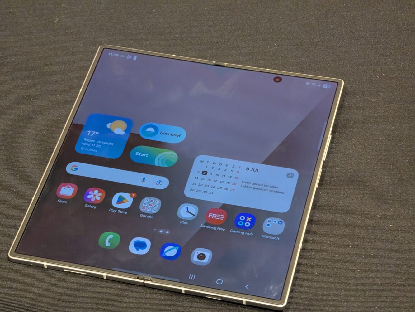 1000027088 Een opvouwbare smartphone van Samsung met een geopend scherm waarop verschillende app-pictogrammen en widgets te zien zijn, rust op een donker oppervlak met textuur.