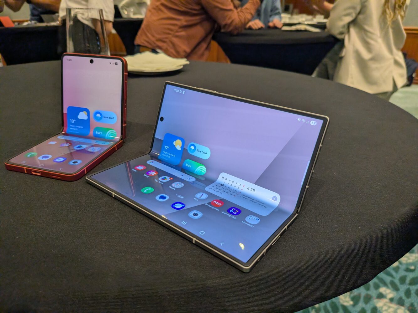 1000027093 Twee opvouwbare smartphones van Samsung worden getoond op een ronde zwarte tafel, elk gedeeltelijk opgevouwen om hun scherm en app-pictogrammen te onthullen. Op de achtergrond zijn mensen zichtbaar.