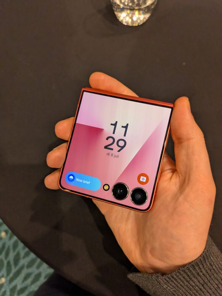 1000027095 Een hand die een opgevouwen Samsung flip smartphone vasthoudt met de tijd en datum op het scherm, met dubbele camera's zichtbaar aan de buitenkant.