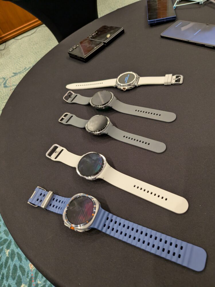 1000027098 Vijf Samsung smartwatches met verschillende gekleurde bandjes liggen op een tafel naast twee smartphones.