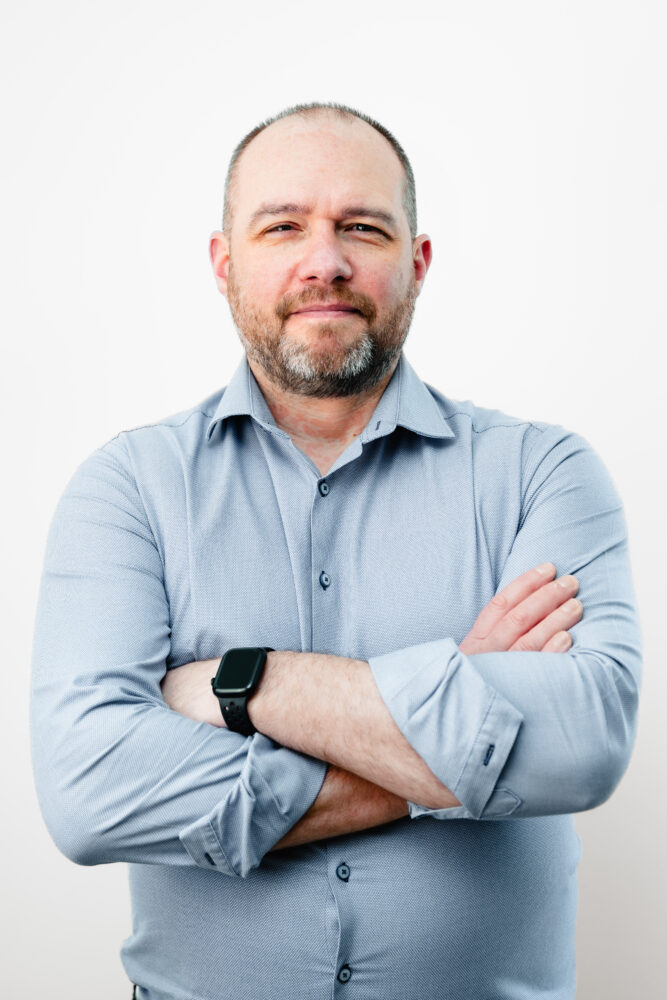 Allan Carchrie – Darktrace Een man van middelbare leeftijd met een baard en kort haar, die een lichtblauw overhemd en een zwarte smartwatch draagt, staat met zijn armen over elkaar tegen een effen witte achtergrond.