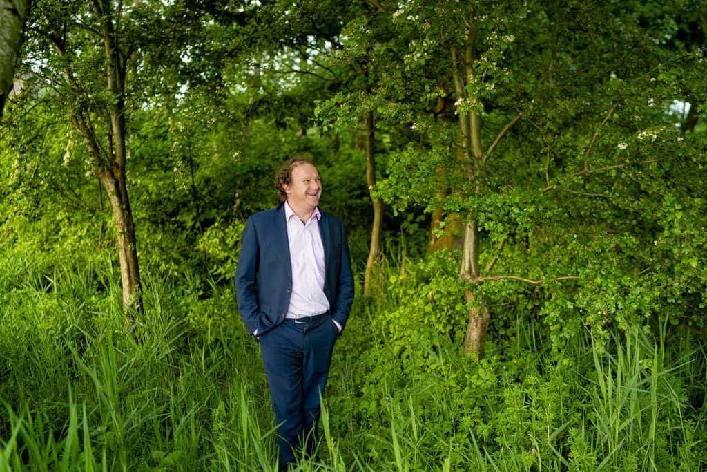 Brenno de Winter Een man in pak staat met zijn handen in zijn zakken te glimlachen terwijl hij door een weelderig groen bebost gebied loopt.