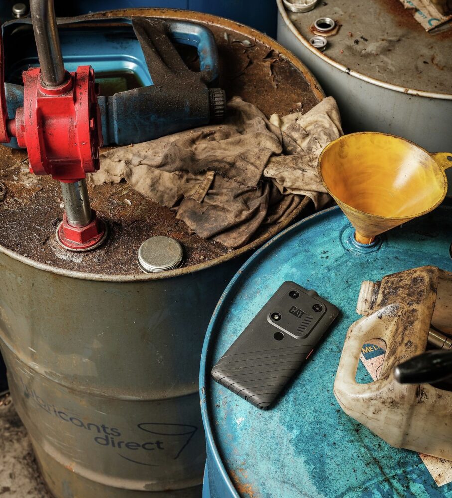 Een robuuste smartphone met een hoge IP-waarde, een vuile doek, een trechter en een pomp worden in een werkplaats bovenop besmeurde industriële vaten geplaatst.