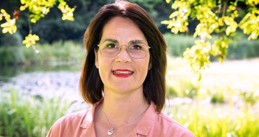 EefjeCuppen_twitter Een vrouw met schouderlang bruin haar en een bril staat buiten bij groen en water en draagt een lichtroze shirt en een halsketting.
