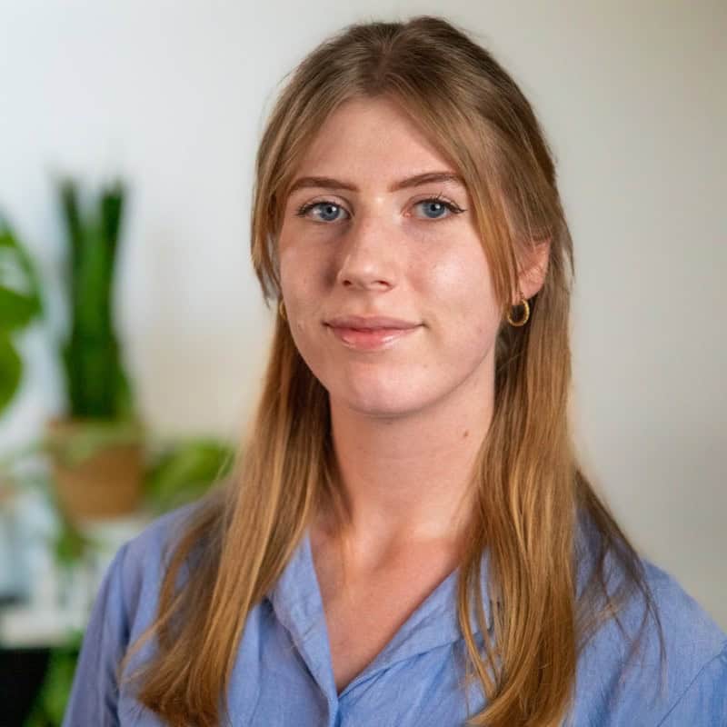 Een jonge vrouw met lang, lichtbruin haar en blauwe ogen, die een lichtblauw shirt draagt, staat binnen met groene planten op de achtergrond.