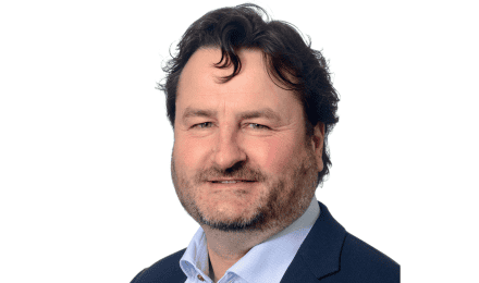 Een man met golvend bruin haar, lichte huid en baard, die een donker colbert en lichtblauw overhemd draagt, poseert tegen een witte achtergrond.