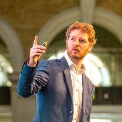 Robert McArdle – Trend Micro Een man met krullend haar en een baard, die een colbert en wit overhemd draagt, houdt een apparaat vast en gebaart met zijn rechterhand terwijl hij binnenshuis spreekt.