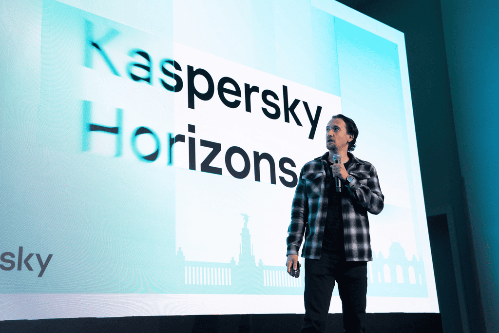 SergeyLozhkin 1 Een persoon houdt een microfoon vast en staat voor een groot scherm met de tekst "Kaspersky Horizons" tijdens een presentatie of evenement.