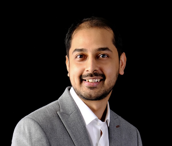 Sudeep Singh_Zscaler ThreatLabz Research Een man met kort zwart haar en een getrimde baard, die een lichtgrijs colbert en wit overhemd draagt, glimlacht tegen een zwarte achtergrond.