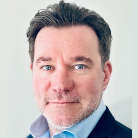 Een man met een lichte huid, blauwe ogen en kort bruin haar, die een lichtblauw overhemd en een donkere blazer draagt, poseert voor een effen, lichte achtergrond.