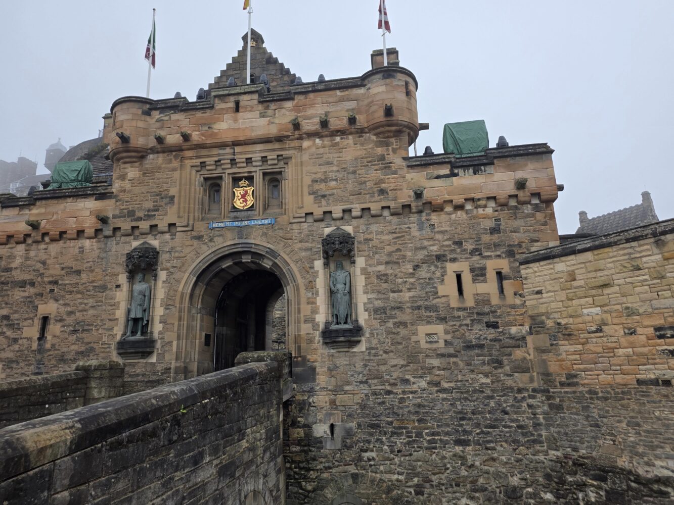 Stenen ingang van een historisch kasteel met standbeelden die de boog flankeren, een wapenschild boven de deur, vlaggen bovenop en een bewolkte lucht op de achtergrond - vastgelegd met de scherpe details van een Samsung Galaxy Z Fold 7.