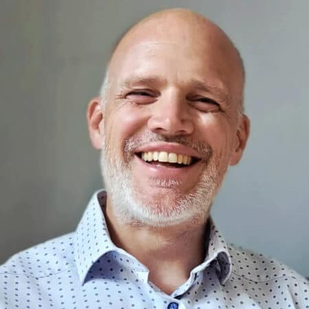 Een man van middelbare leeftijd met een kaal hoofd en een korte witte baard glimlacht. Hij draagt een lichtblauw shirt met patroon en staat voor een effen grijze achtergrond.