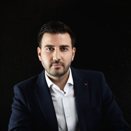 Een man met kort donker haar en een baard, die een marineblazer, wit overhemd en blauwe spijkerbroek draagt, zit in kleermakerszit tegen een zwarte achtergrond en kijkt naar de camera.