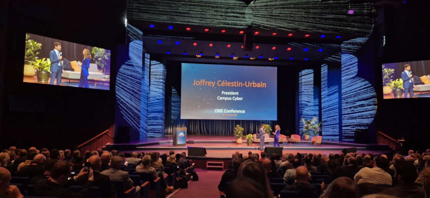 Een spreker staat op het podium van een conferentie met op een scherm "Joffrey Célestin-Urbain, President, Campus Cyber" en een publiek dat in een schemerig verlicht auditorium zit.