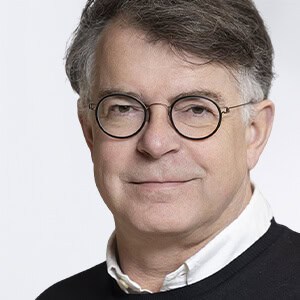 Man van middelbare leeftijd met kort grijs haar, ronde bril en een lichte glimlach, draagt een wit overhemd onder een donkere trui, tegen een effen lichte achtergrond.