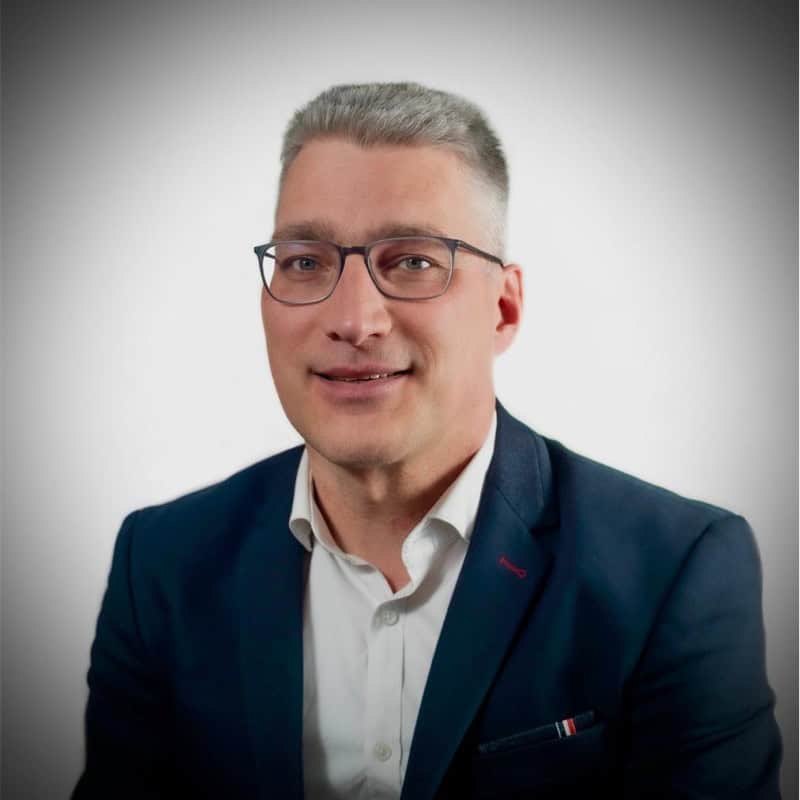 Een man met kort grijs haar en een bril draagt een donker colbert en wit overhemd, lachend voor een effen witte achtergrond.