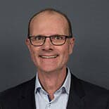Peter van Eck_1 Een man met bril en terugwijkende haarlijn, in een donker colbert en lichtblauw overhemd, glimlacht voor een effen donkere achtergrond.